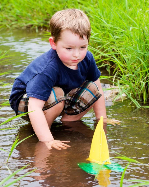 Menino se diverte brincando com barcos de brinquedo em riacho no parque durante a primavera ou verão — Fotografia de Stock