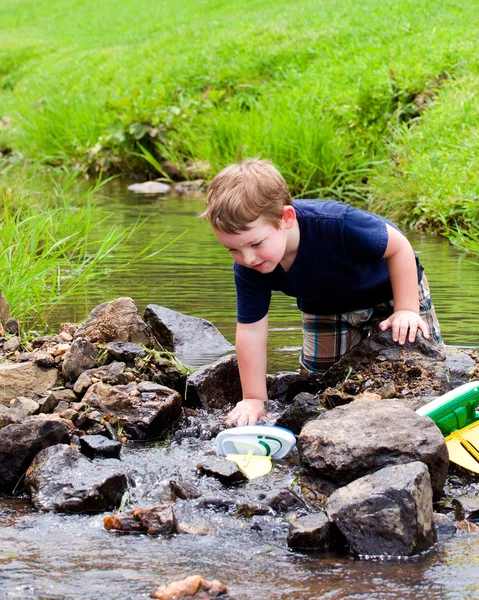 Menino se diverte brincando com barcos de brinquedo em riacho no parque durante a primavera ou verão — Fotografia de Stock