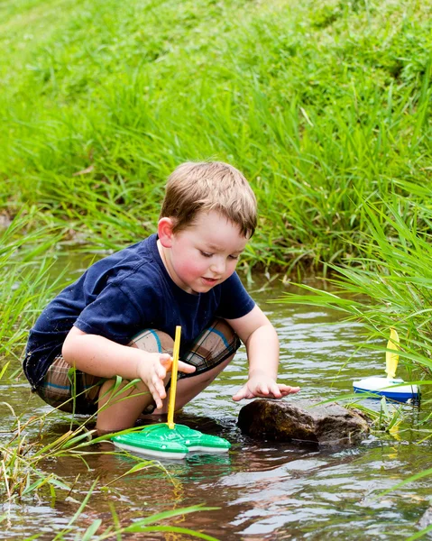 Chico se divierte jugando con barcos de juguete en arroyo en el parque durante la primavera o el verano —  Fotos de Stock