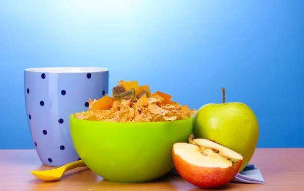 Flocos de milho saborosos em tigela verde, maçãs e vidro de leite na mesa de madeira no fundo azul — Fotografia de Stock
