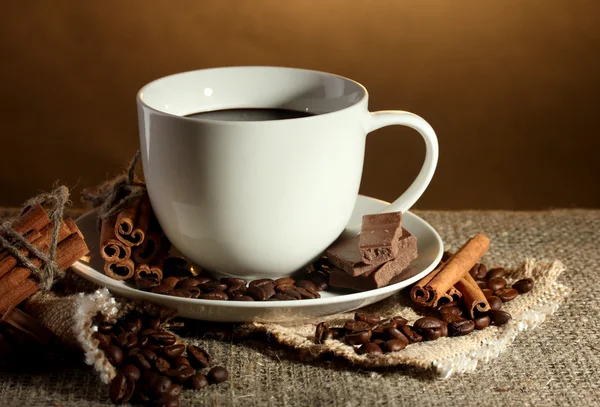Secangkir kopi dan kacang-kacangan, kayu manis tongkat dan coklat pada karung di latar belakang coklat — Stok Foto