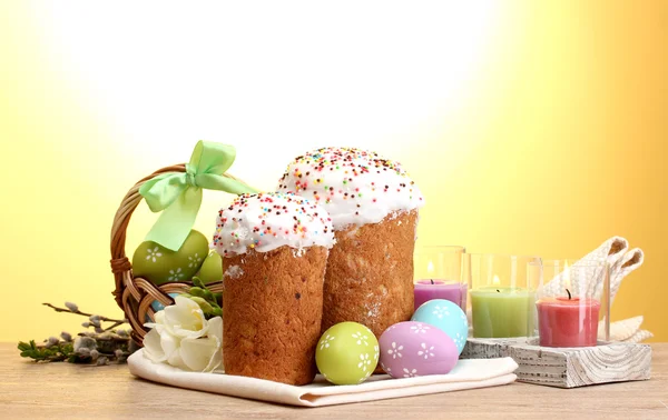Красиві великодні торти, барвисті яйця в кошику і свічки на дерев'яному столі на жовтому тлі — стокове фото