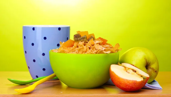 Tasty cornflakes in green bowl, apples and glass of milk on wooden table on green background — Stock Photo, Image
