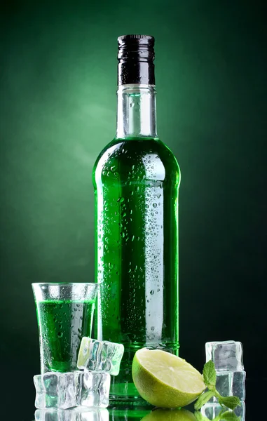 Bottiglia e bicchiere di assenzio con calce e ghiaccio su fondo verde — Foto Stock