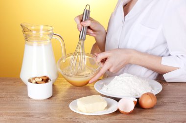 Female hands mixing eggs in bowl on wooden table on yellow background clipart