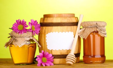 Sweet honey in jars and barrel with drizzler on wooden table on green background