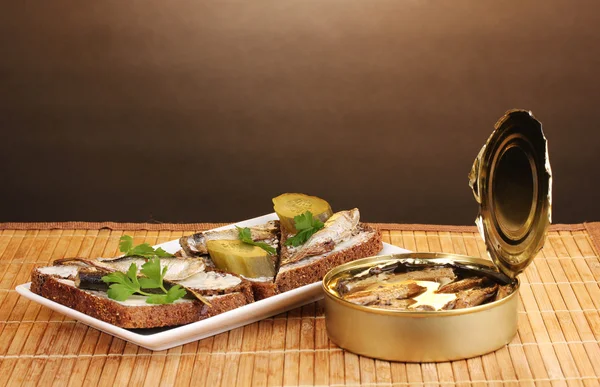 Tasty sandwiches with sprats on plate on wooden mat on brown background — Stock Photo, Image