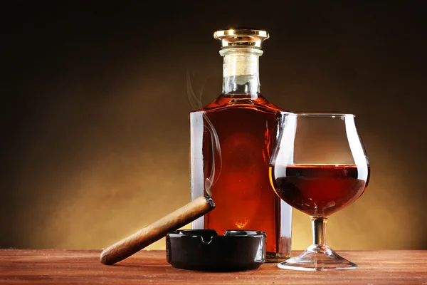 Fles en glas brandy en sigaar op houten tafel op bruine achtergrond — Stockfoto