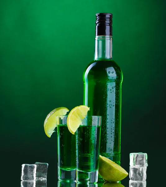 Bottle and glasses of absinthe with lime and ice on green background — Stock Photo, Image