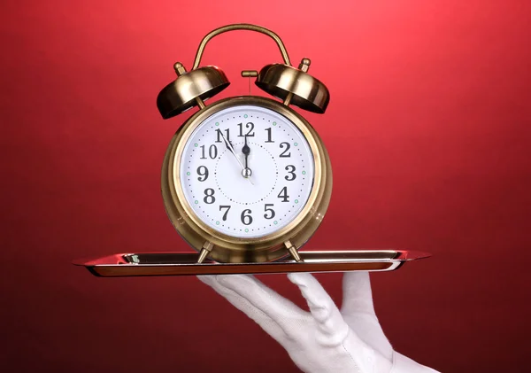 Hand in glove holding silver tray with alarm clock on red background — Stock Photo, Image