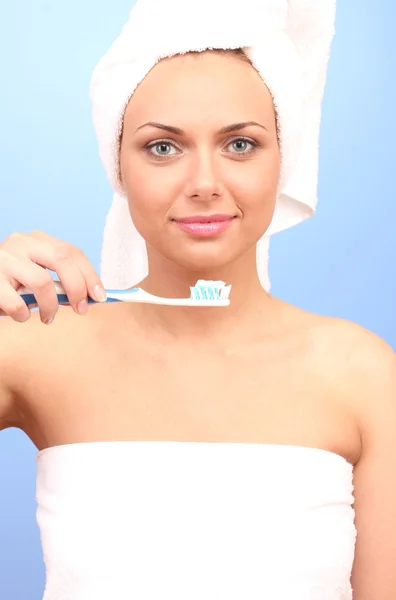 Schöne junge Frau nach der Dusche mit einem Handtuch auf dem Kopf und einer Zahnbürste in der Hand auf blauem Hintergrund in Großaufnahme — Stockfoto