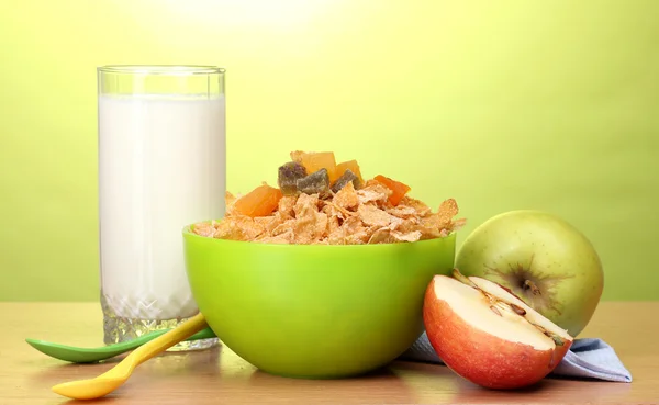 Välsmakande cornflakes i grön skål, äpplen och glas mjölk på träbord på grön bakgrund — Stockfoto