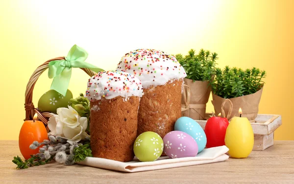 Красивые пасхальные торты, красочные яйца в корзине и свечи на деревянном столе на желтом фоне — стоковое фото