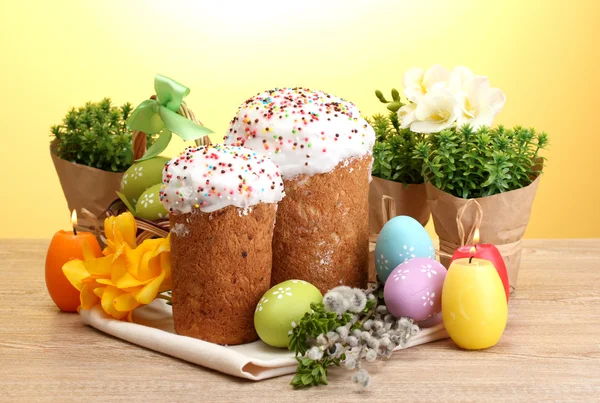 Piękne wielkanocne ciasta, kolorowe jajka i świece na drewnianym stole na żółtym tle — Zdjęcie stockowe