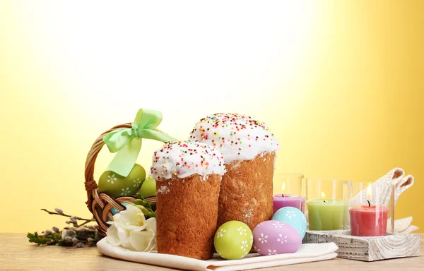 stock image Beautiful Easter cakes, colorful eggs in basket and candles on wooden table on yellow background