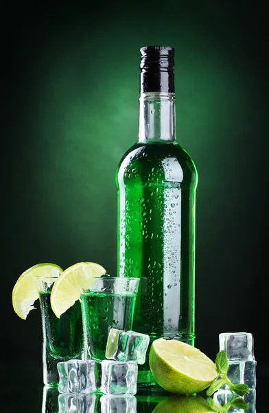 Bottle and glasses of absinthe with lime and ice on green background — Stock Photo, Image