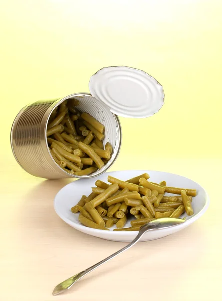 Open tin can and plate with french bean and spoon on wooden table on green background — Stock Photo, Image