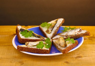 Tasty sandwiches with sprats on plate on wooden table on brown background clipart