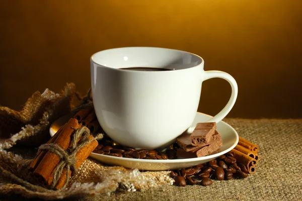Kopp kaffe och bönor, kanelstänger och choklad på säckväv på brun bakgrund — Stockfoto