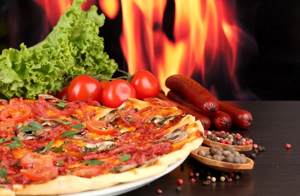 Deliziosa pizza, salumi, pomodori e spezie sul tavolo di legno su sfondo fiamma — Foto Stock