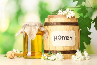 Sweet honey in barrel and jar with acacia flowers on wooden table on green background clipart