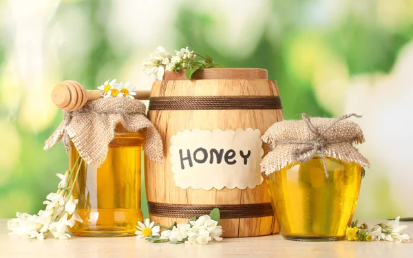 Édes méz hordó és üvegekbe a virágok akác fából készült asztal zöld háttér — Stock Fotó