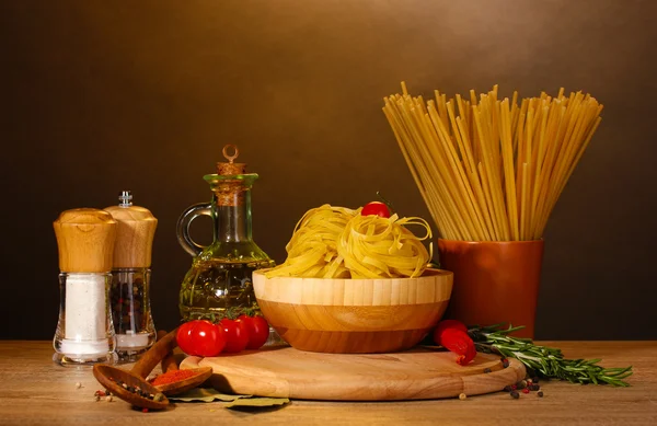 Spaghetti, nudlar i skål, burk med olja och grönsaker på träbord på brun bakgrund — Stockfoto