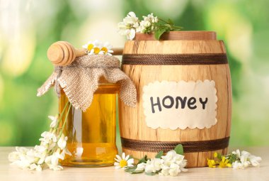 Sweet honey in barrel and jar with acacia flowers on wooden table on green background clipart