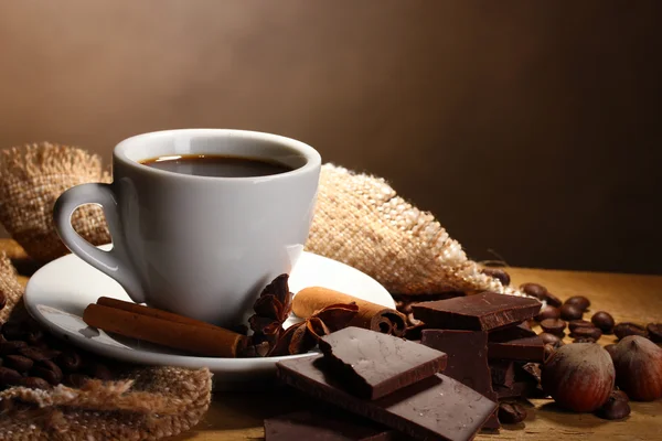 Taza de café y frijoles, palitos de canela, nueces y chocolate en la mesa de madera sobre fondo marrón —  Fotos de Stock