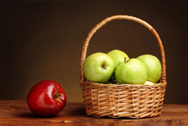 Manzanas verdes jugosas en cesta y manzana roja sobre mesa de madera sobre fondo marrón —  Fotos de Stock