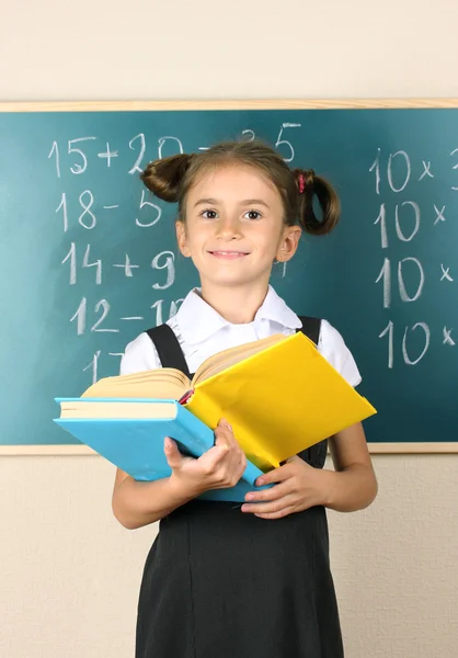 Beautiful little girl with books standing near blackboard in classroom — Stock Photo, Image