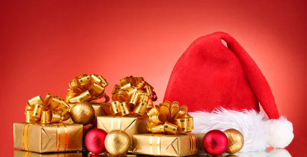 Schöne Weihnachtsmütze, Geschenke und Weihnachtskugeln auf rotem Hintergrund — Stockfoto