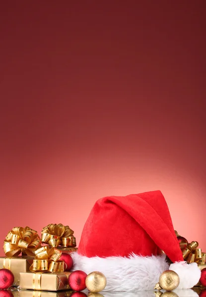 Mooie kerstmuts, giften en kerstballen op rode achtergrond — Stockfoto