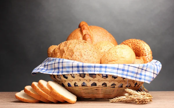 Delicious bread in basket and ears on wooden table on gray background — Stock Photo, Image