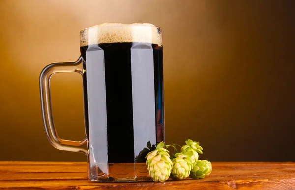 Dunkles Bier im Becher und grüner Hopfen auf Holztisch auf braunem Hintergrund — Stockfoto