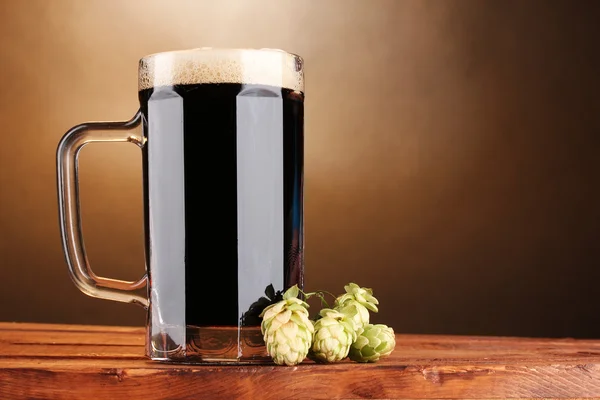 Tmavé pivo v hrnek a zelené hop na dřevěný stůl na hnědé pozadí — Stock fotografie