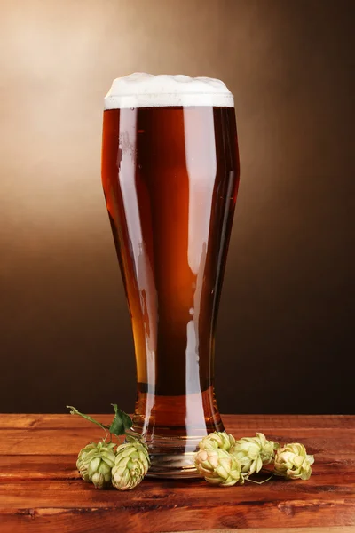 Пиво в стекле и зеленый хмель на деревянном столе на коричневом фоне — стоковое фото