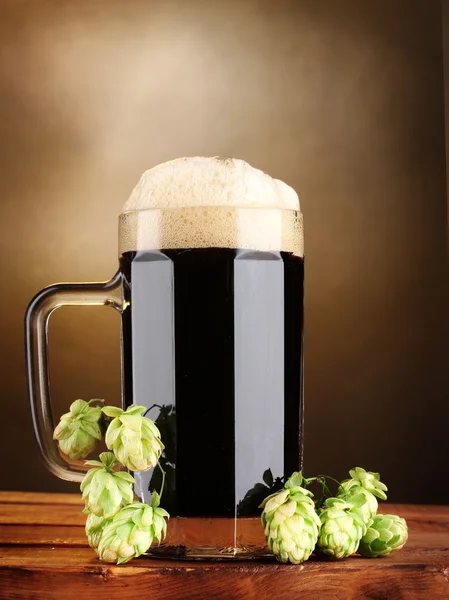 Dunkles Bier im Becher und grüner Hopfen auf Holztisch auf braunem Hintergrund — Stockfoto