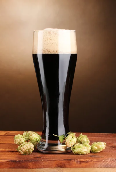 Donker bier in glas en groen hop op houten tafel op bruine achtergrond — Stockfoto