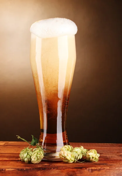 Пиво в стекле и зеленый хмель на деревянном столе на коричневом фоне — стоковое фото