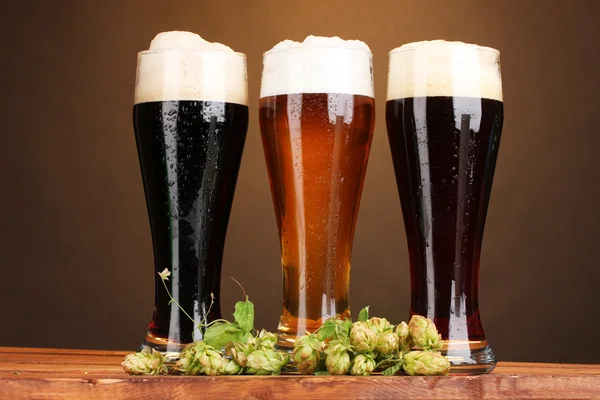Три келихи з різними пивом і хоп на дерев'яному столі на коричневому фоні — стокове фото