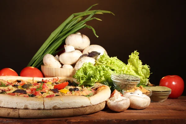 Delicious pizza, vegetables and spices on wooden table on brown background — Stock Photo, Image