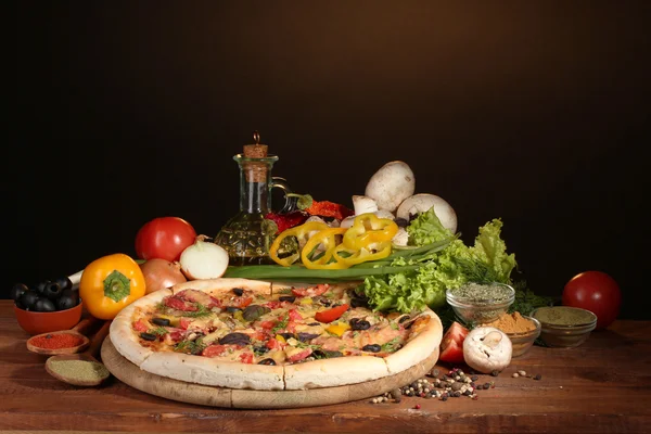 Herkullinen pizza, vihannekset ja mausteet puupöydällä ruskealla taustalla — kuvapankkivalokuva