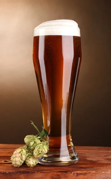 Cerveza en vidrio y lúpulo verde sobre mesa de madera sobre fondo marrón —  Fotos de Stock