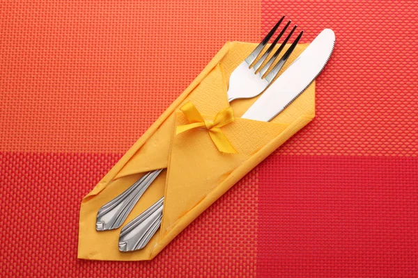 Fork and knife in a yellow cloth with a bow on a red tablecloth — Stock Photo, Image