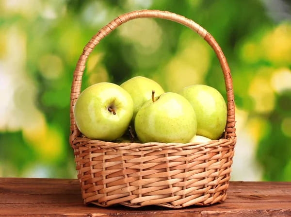 Saftigt gröna äpplen i korgen på träbord på grön bakgrund — Stockfoto