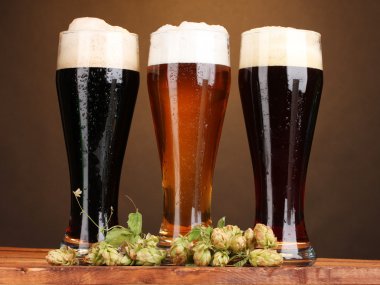 Three glasses with different beers and hop on wooden table on brown backgro clipart