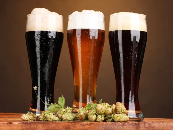 Three glasses with different beers and hop on wooden table on brown backgro — Stock Photo, Image