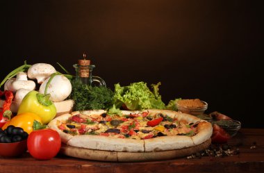 Delicious pizza, vegetables and spices on wooden table on brown background clipart