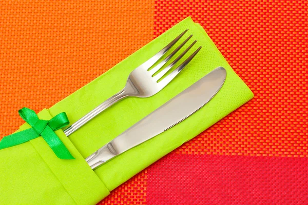 Fork and knife in a green cloth with a bow on a red tablecloth — Stock Photo, Image